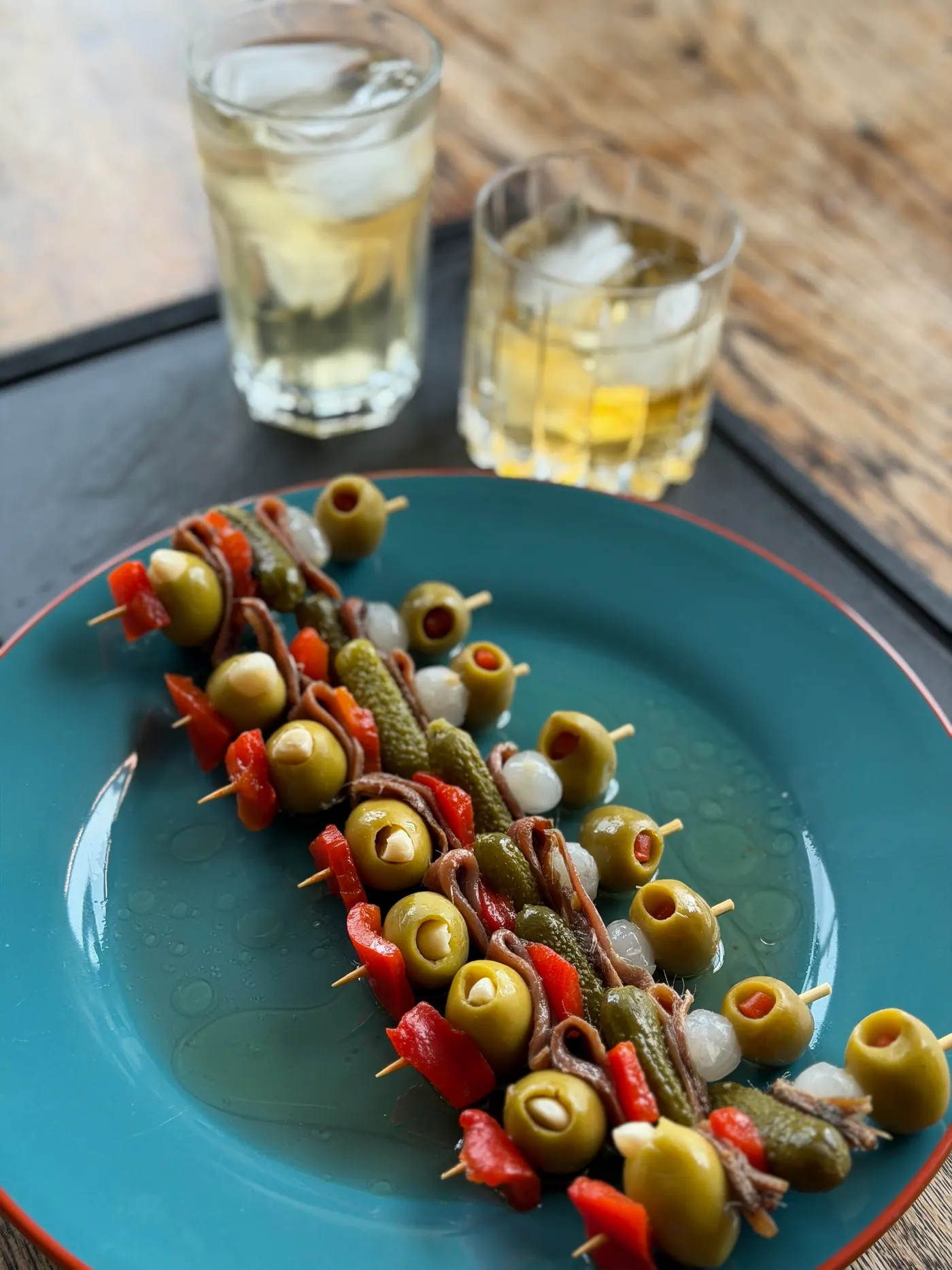 A row of olive and pepper skewers on a blue plate beside two glasses of vermouth on ice