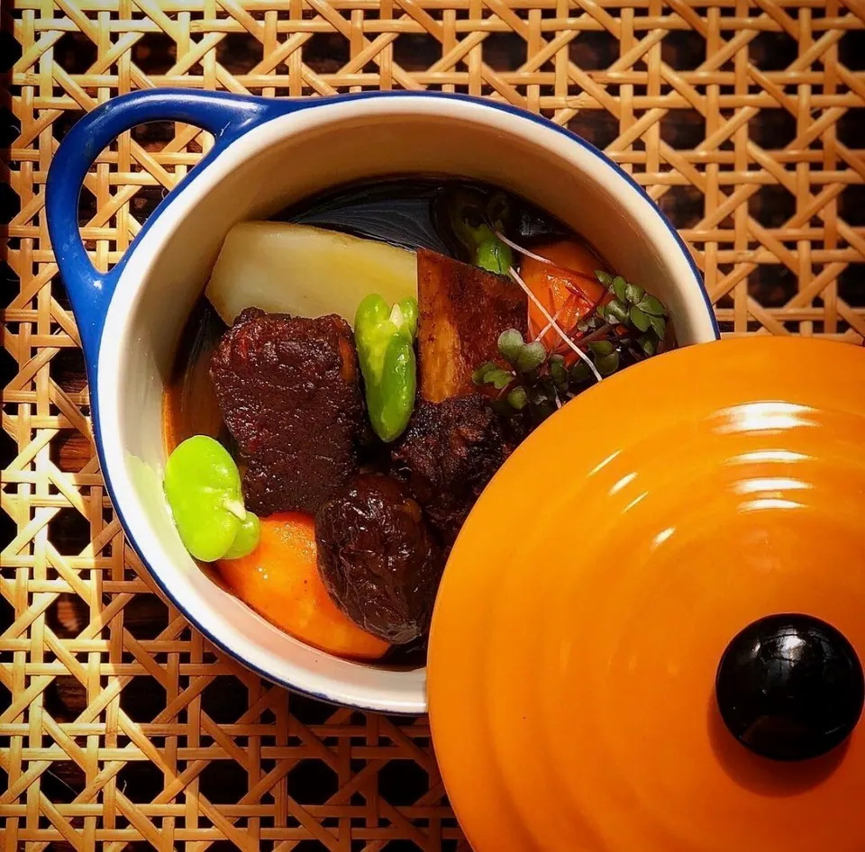 A braise of beef, potatoes and peppers in a small blue pot beside its orange lid